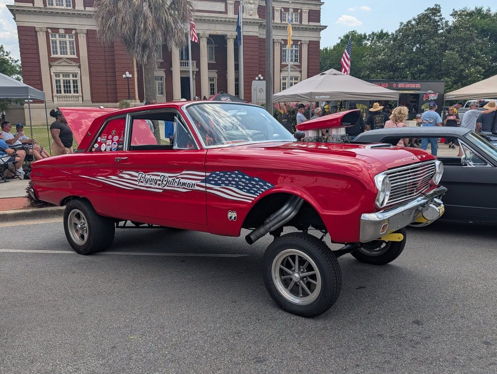 Bright red vintage drag racing car with an American flag motif, elevated front, and &ldquo;Flying Dutchman&rdquo; on its side, displayed at an outdoor car show.