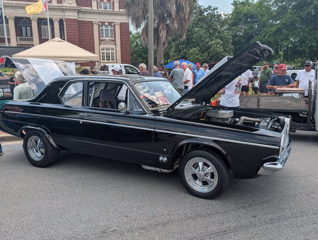 A classic black car with its hood open on display at a car show, surrounded by people and tents, parked on a street in front of a brick building.