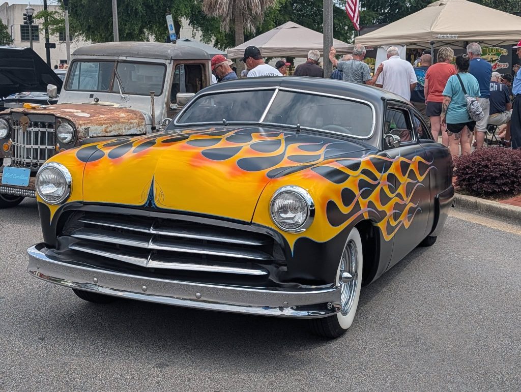 Classic car with yellow and orange flame paint job displayed at a car show, surrounded by people and other vintage vehicles.