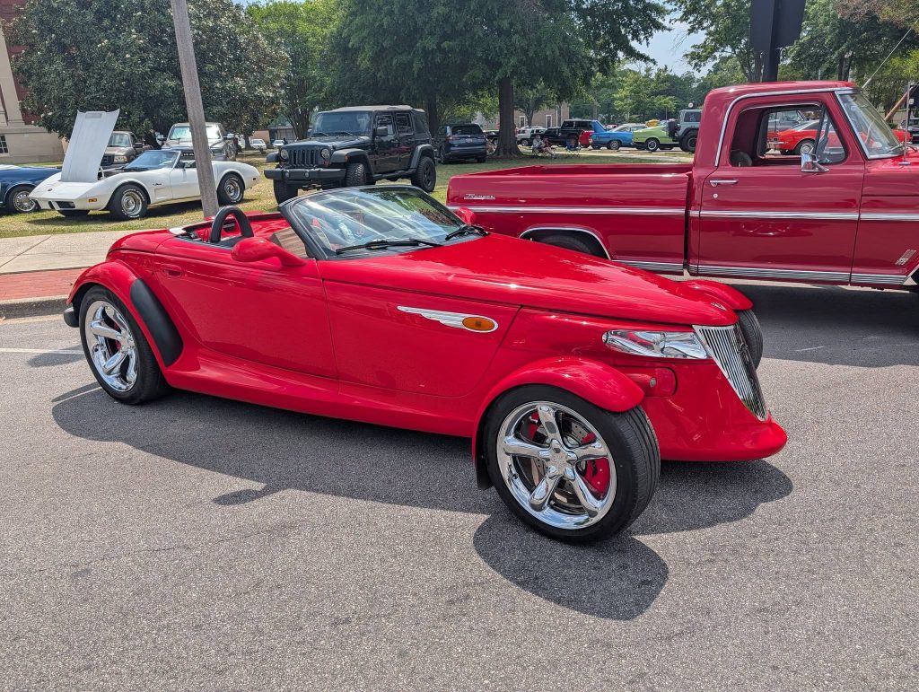 A bright red convertible sports car parked at a car show, with other classic vehicles visible in the background on a sunny day.