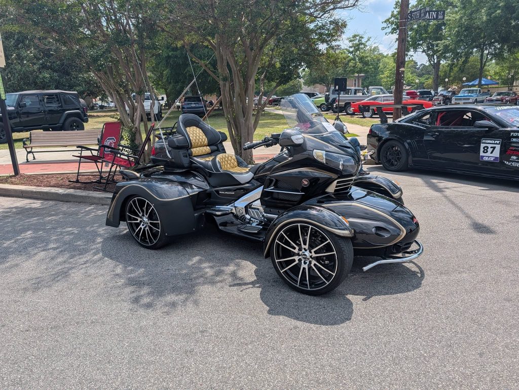 A black and gold three-wheeled motorcycle with large rims is parked on a street at a car show, surrounded by other vehicles and outdoor seating.