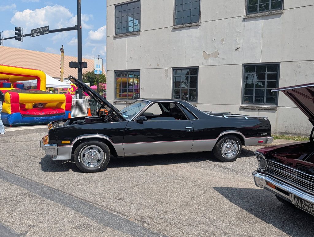 A black classic car with its hood open is parked on a street near a bounce house and a building with multiple windows.
