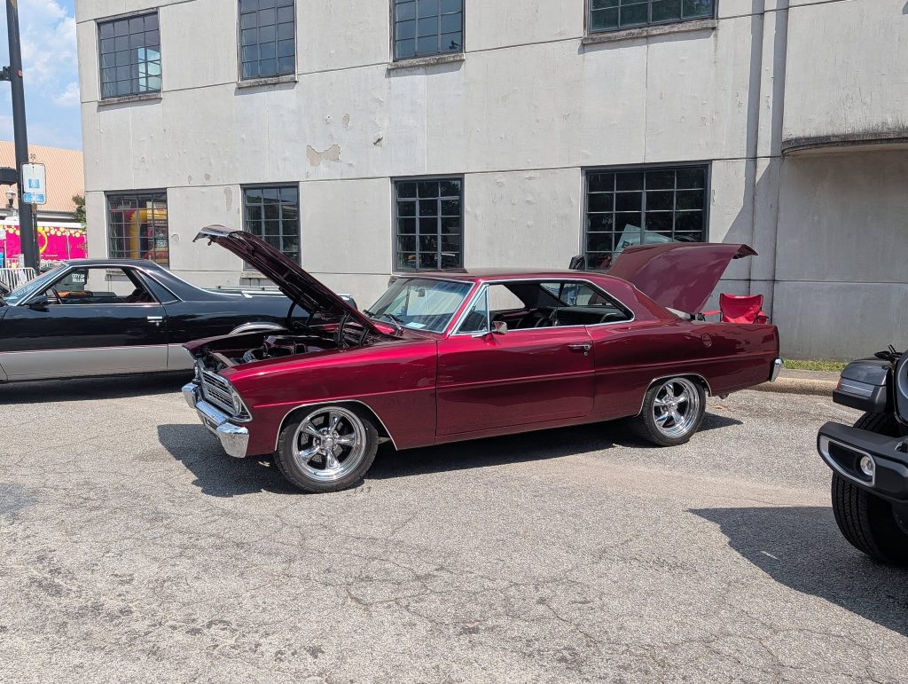 A classic maroon muscle car with its hood and trunk open is parked at a car show, next to other vintage vehicles, in front of a gray building.