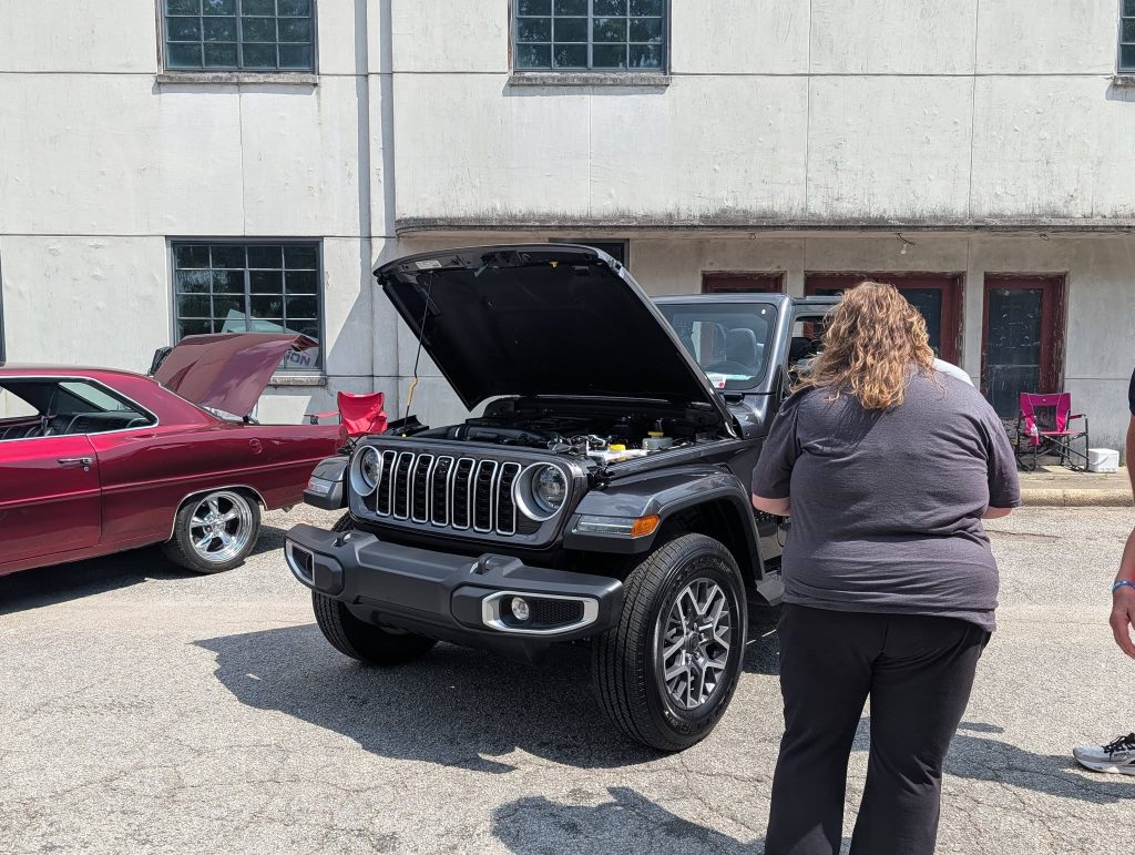 A person stands near a black SUV with its hood open at a car show, with another red classic car beside it.