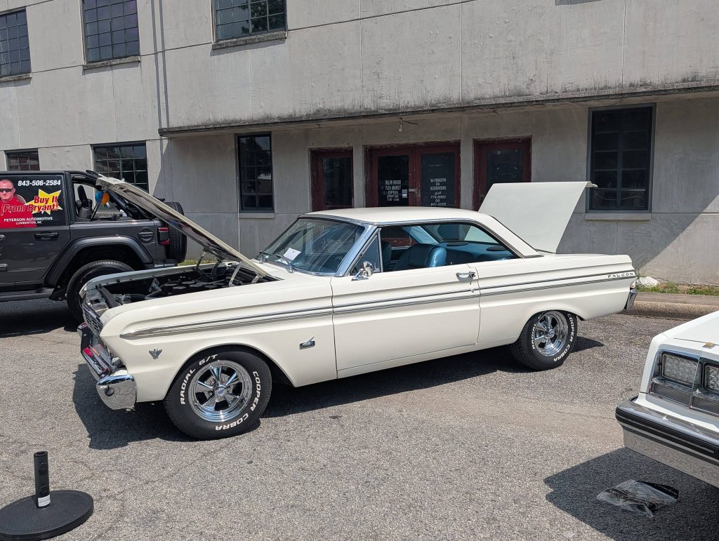 A classic white Ford Falcon car on display outdoors with its hood and trunk open, parked beside other vehicles.