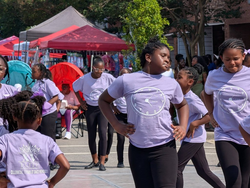 Young girls in matching purple shirts and black pants perform a group dance at an outdoor event, with tents and onlookers in the background.