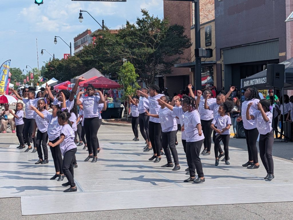 A group of children in matching outfits perform a choreographed dance routine outdoors at a street festival with a crowd watching.
