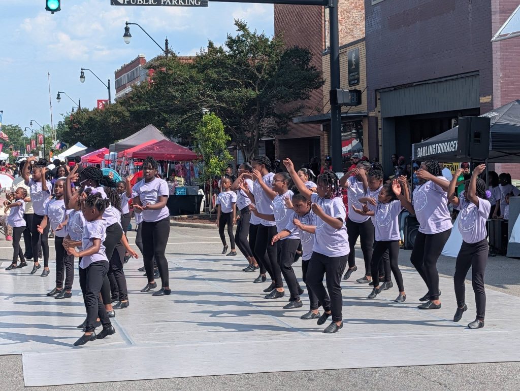 A group of children in matching outfits performs a dance routine outdoors on a city street during a public event or festival.