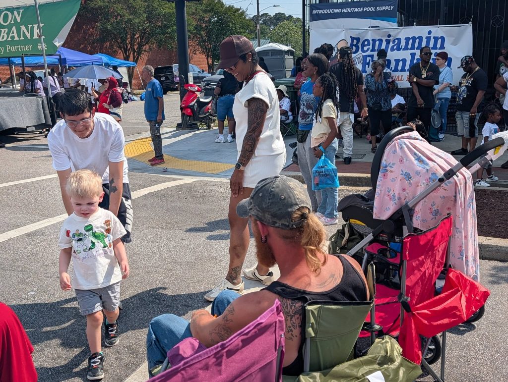 People gather outdoors at a street event, including children, adults, a stroller, and lawn chairs, with banners and tents visible in the background.