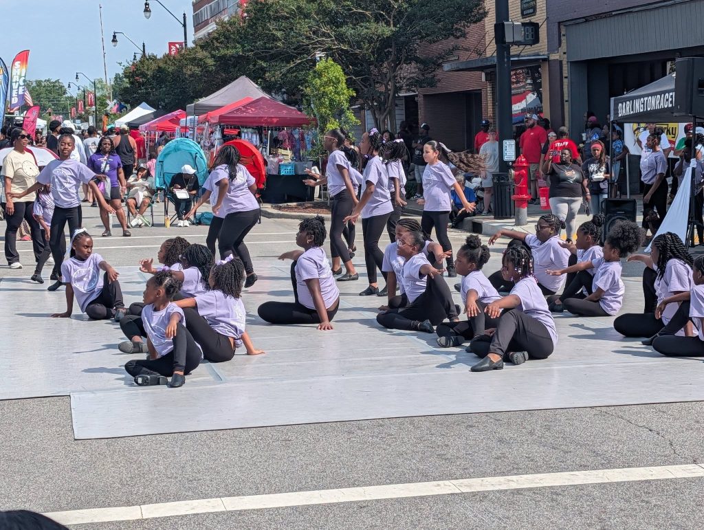 A group of children in matching outfits perform a dance routine on an outdoor stage at a street festival, with spectators and vendor tents in the background.