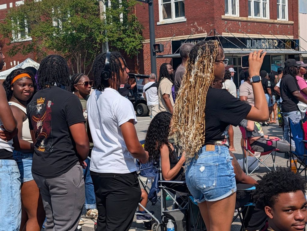 A group of people gathered outdoors at a street event, some standing, some sitting in chairs; one person is recording with a smartphone.