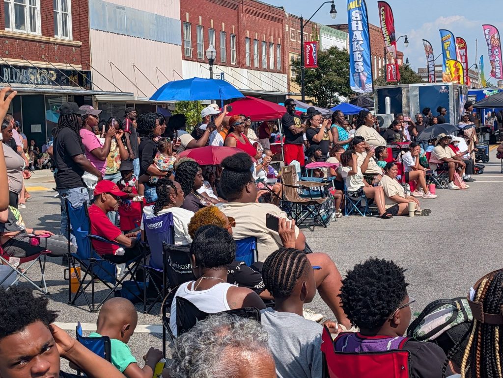 A large crowd, many seated in folding chairs and some holding umbrellas, gathers outdoors at a street festival under sunny skies.