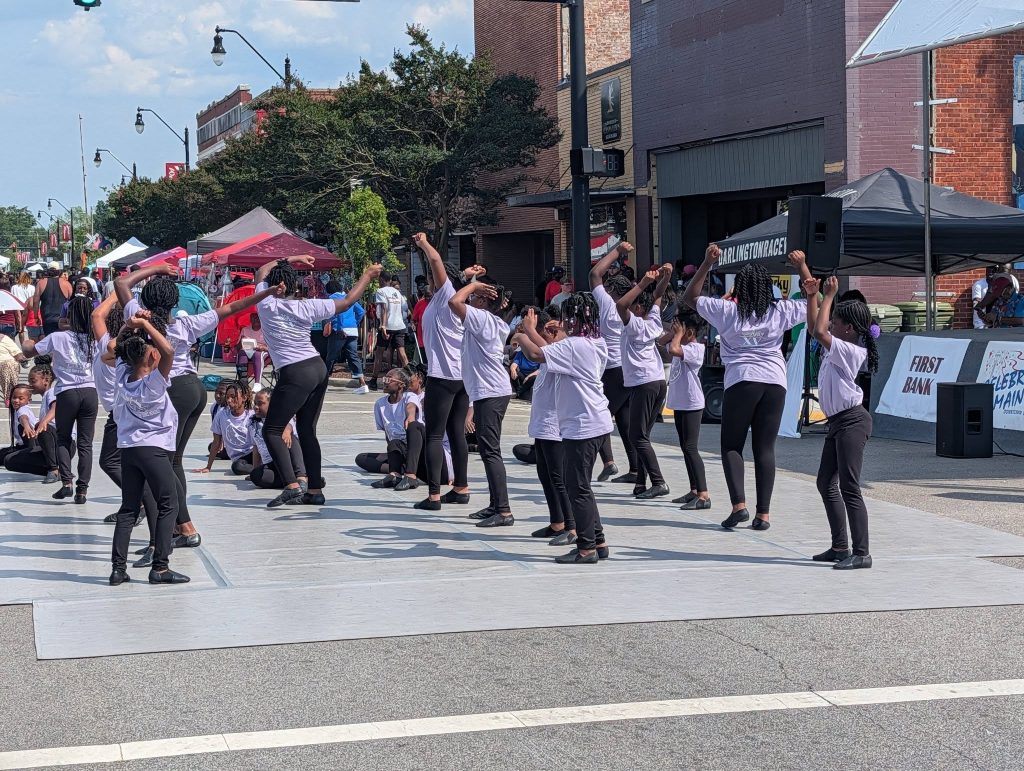 A group of children in matching purple shirts and black pants perform a dance routine outdoors on a city street during a festival or event.