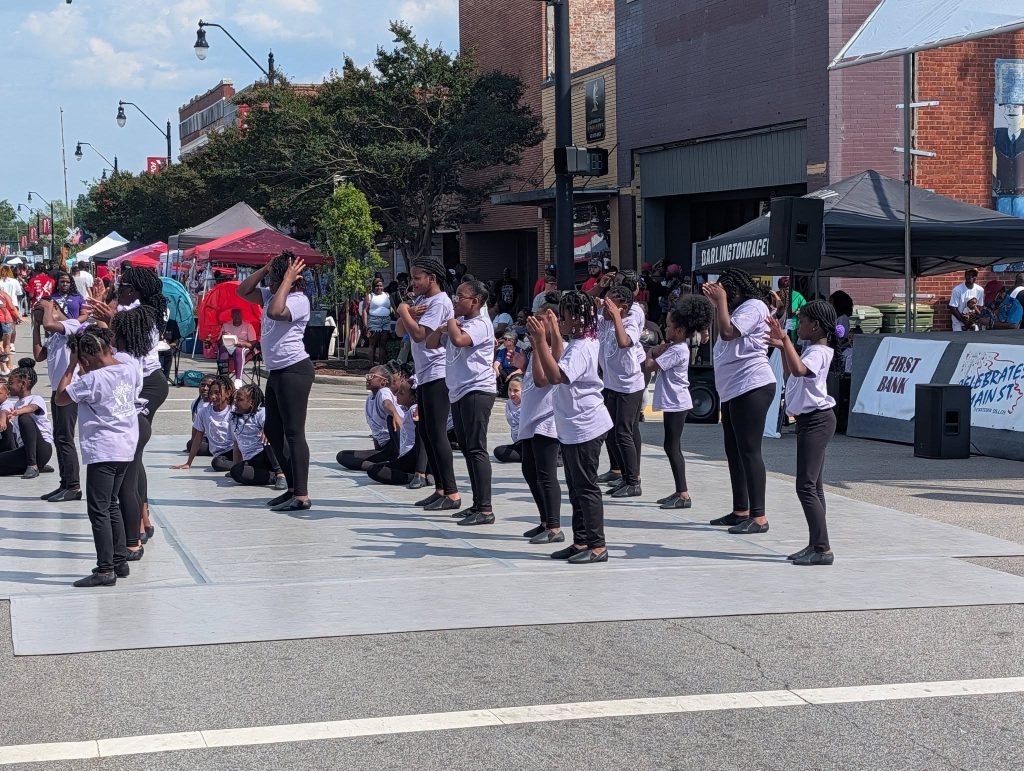A group of children in matching outfits perform a dance routine on an outdoor stage during a street festival.