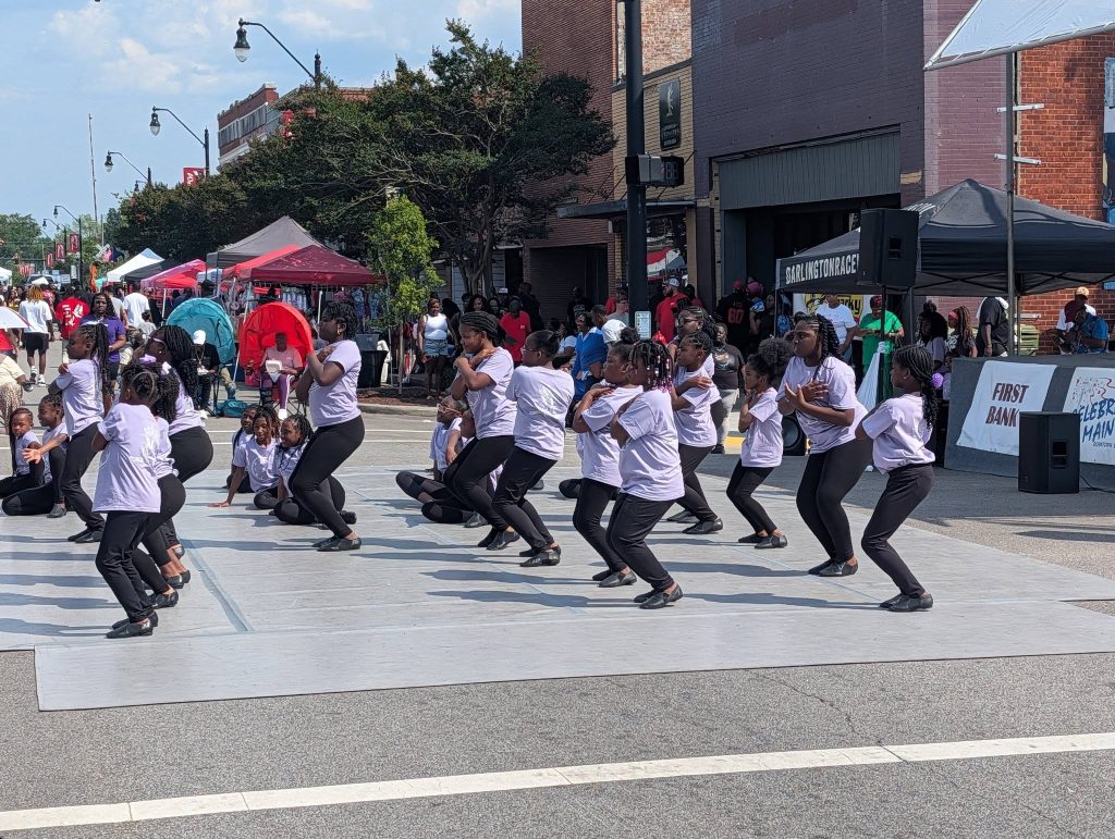 A group of young girls in matching outfits perform a dance routine outdoors on a street during a community event.