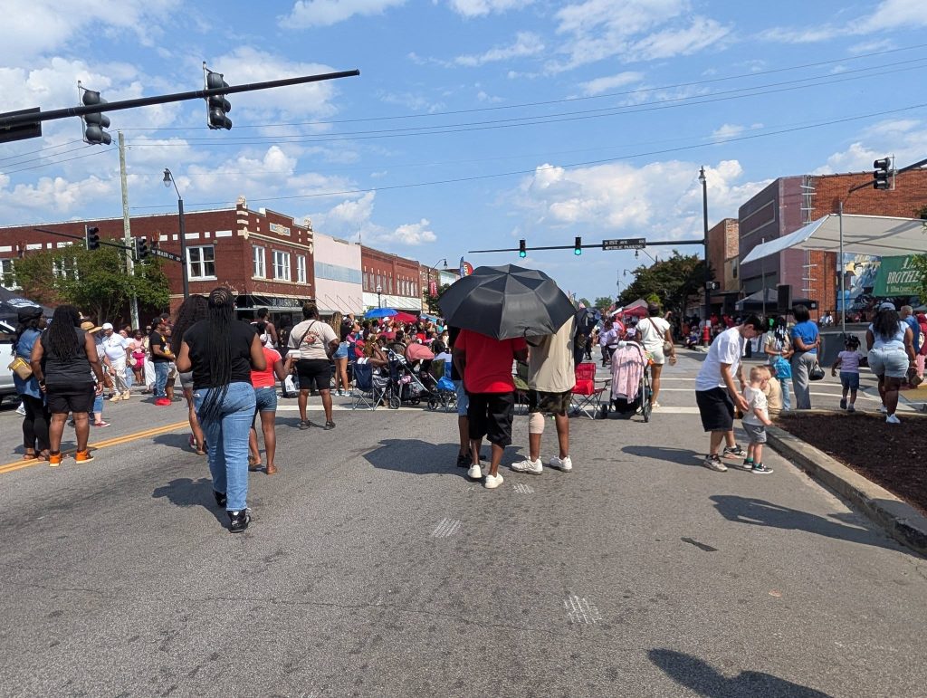 Crowd gathers on a downtown street for a festival or parade, some people sit, others walk, and a few use umbrellas for shade under a sunny sky.