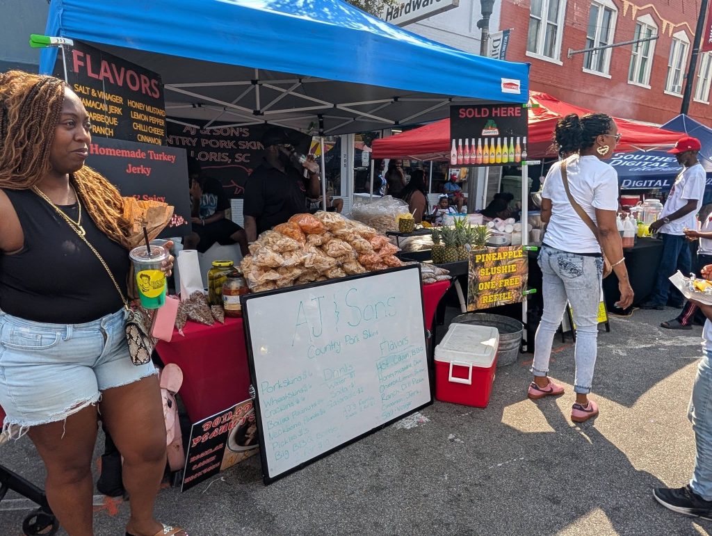People browsing and buying snacks at an outdoor street market, featuring a booth selling pork skins, drinks, and flavored snacks under a blue canopy.