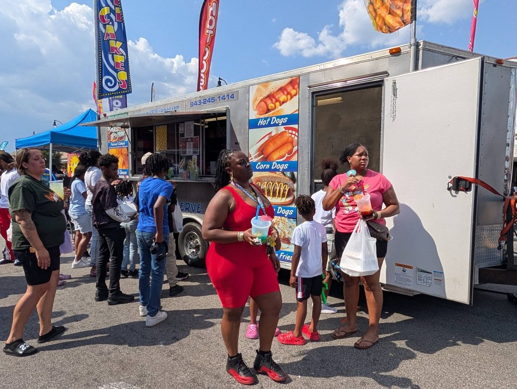 People wait in line at a food truck selling hot dogs, corn dogs, and drinks on a sunny day at an outdoor event.