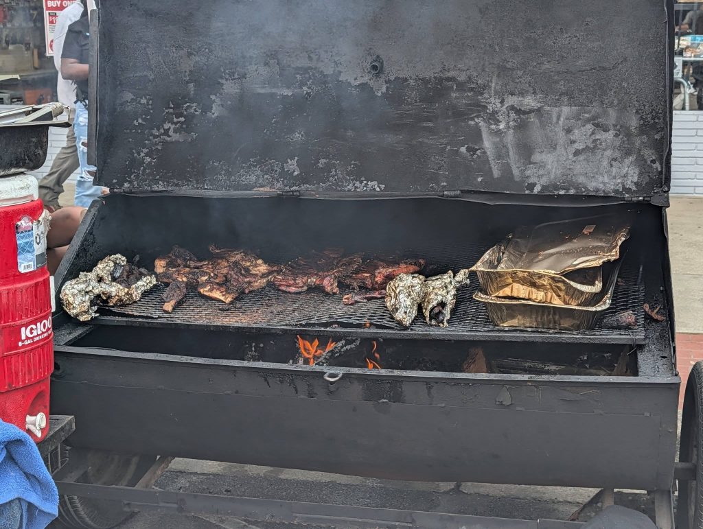 Open barbecue smoker with assorted meats, foil-wrapped food, and trays on the grill, flames visible underneath, with people and a red cooler nearby.