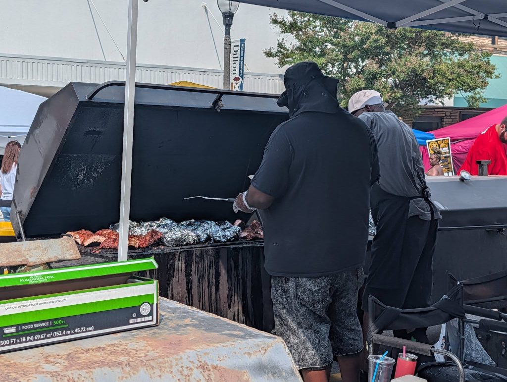 Two people grill meat and foil-wrapped food on a large outdoor barbecue, with boxes and drinks nearby, during a street event or festival.