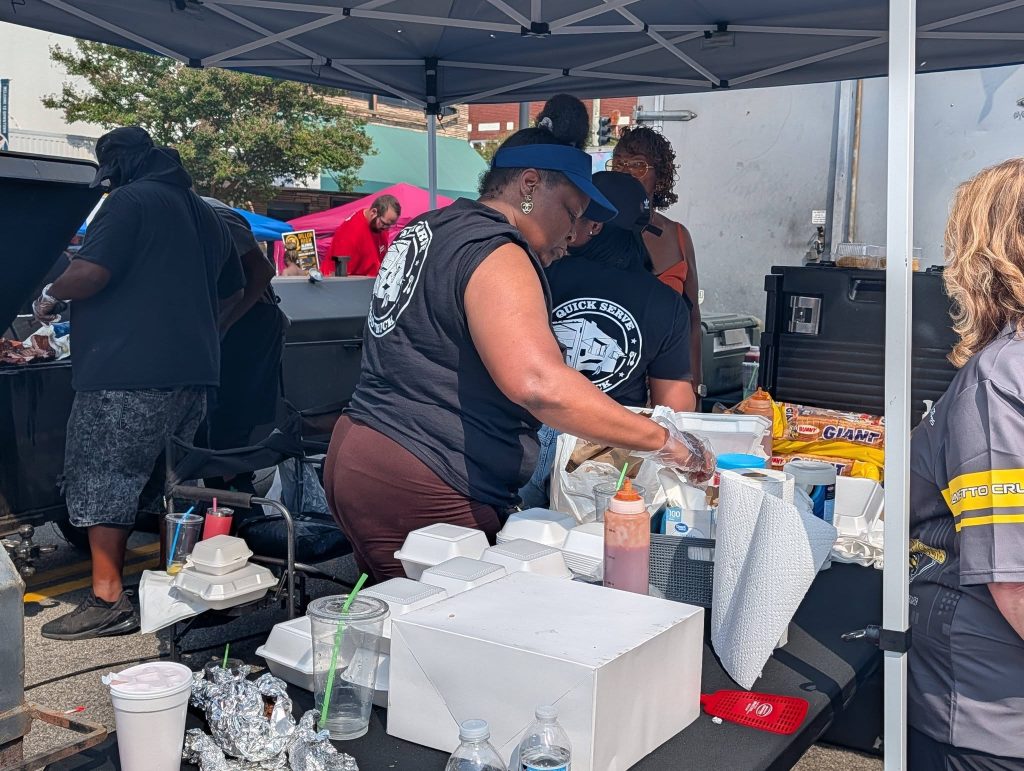 People preparing food at an outdoor event under a tent, with takeout containers, sauces, and supplies on tables, while others grill meat in the background.