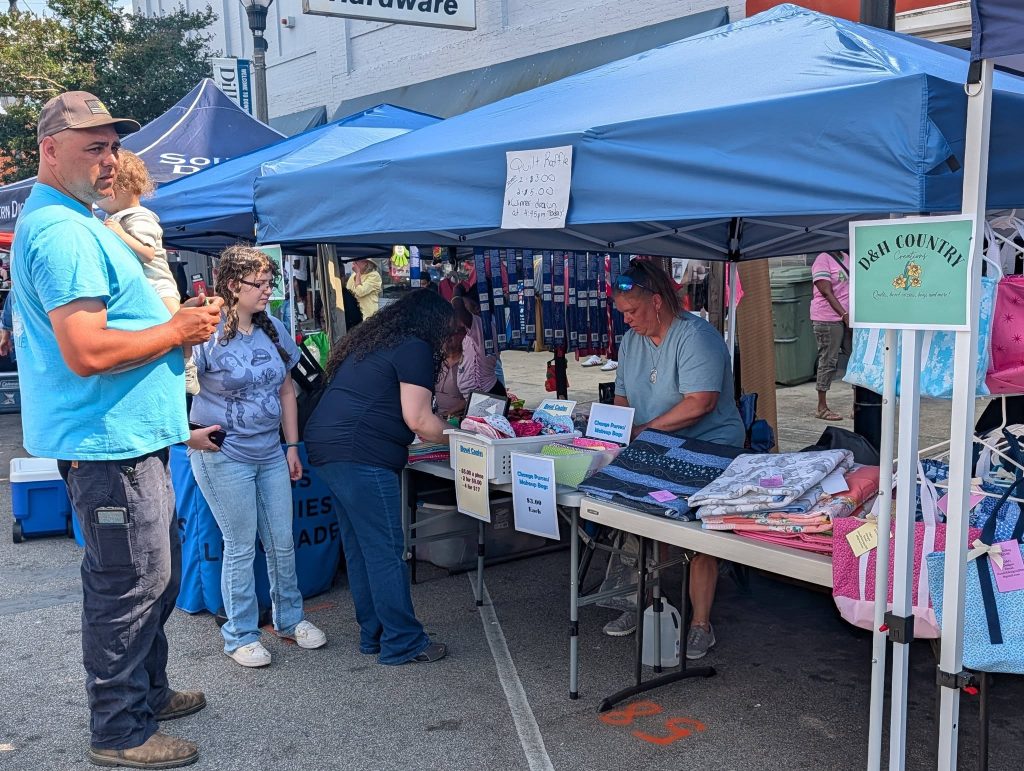 People browsing a craft booth with quilts, bags, and handmade items under a blue tent at an outdoor market.