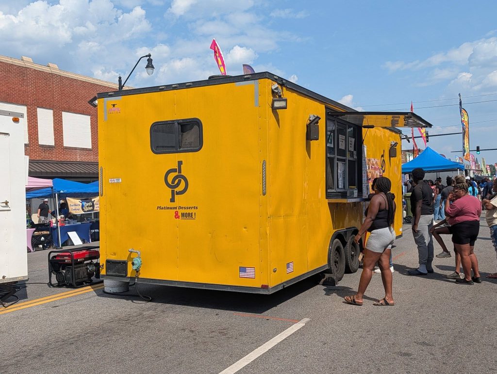 People standing in line at a bright yellow dessert food truck during a street festival, with tents and crowds in the background.