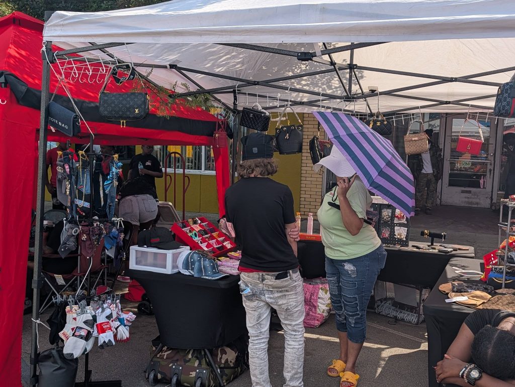 People browsing a street market stall selling handbags, accessories, and socks. A woman holds a purple striped umbrella while talking to another person under the tent.