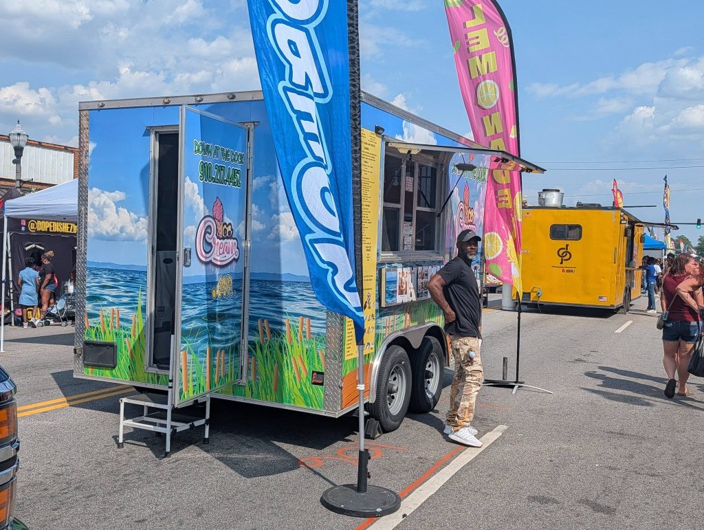 Food truck with colorful beach-themed design and lemonade flags at a street fair, people walking and gathering around in sunny weather.