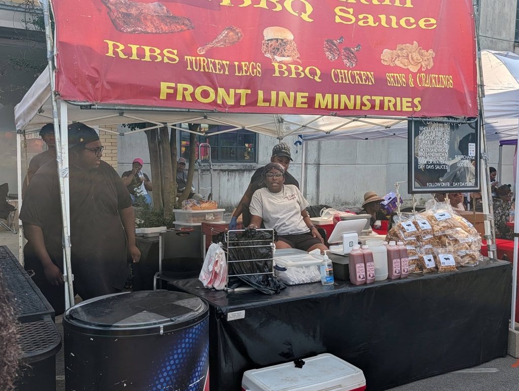 Outdoor BBQ food stall with staff, a large sign advertising ribs, turkey legs, and chicken, bottles of sauce, and bags of pork cracklings on display.