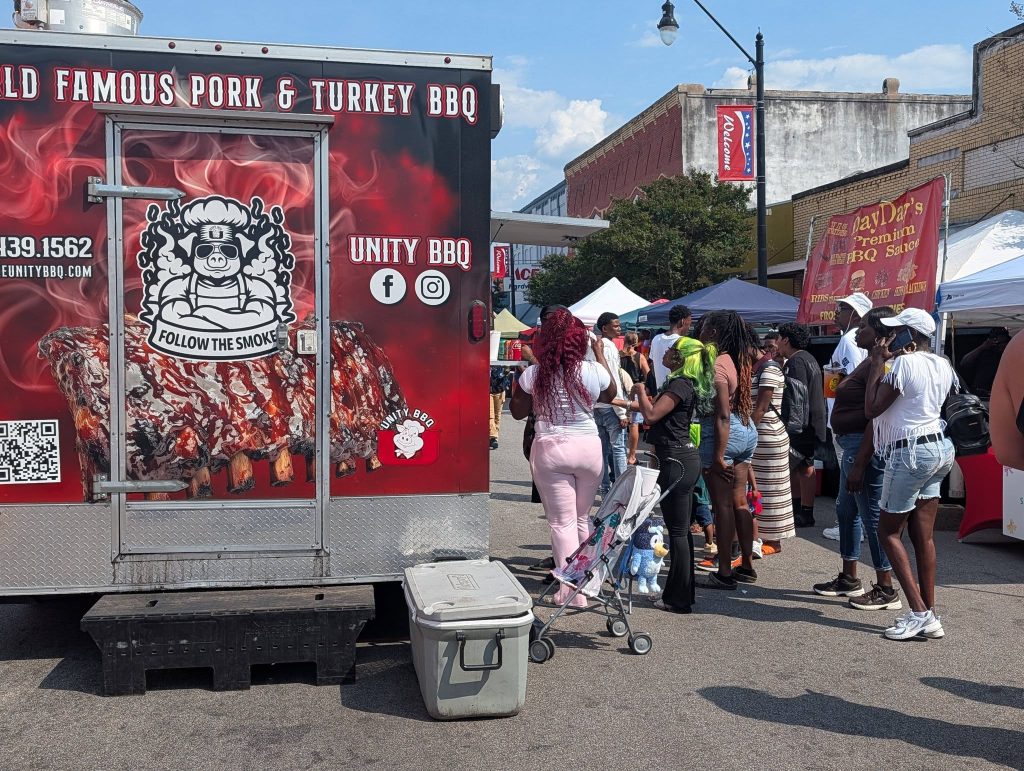 People wait in line at a BBQ food truck, with a colorful &ldquo;Follow the Smoke&rdquo; logo, during an outdoor street festival on a sunny day.