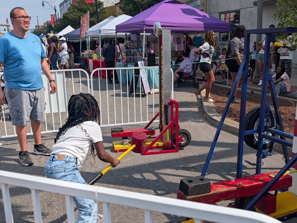 A child tries a hammer strength game at an outdoor street fair, surrounded by people, vendor tents, and festival decorations.