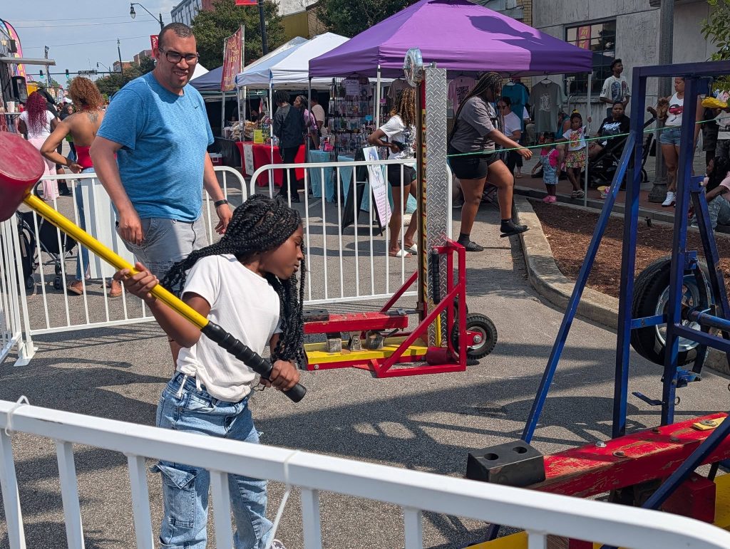 A young girl plays a carnival strength game with a mallet at an outdoor fair, while people watch and shop at market booths in the background.