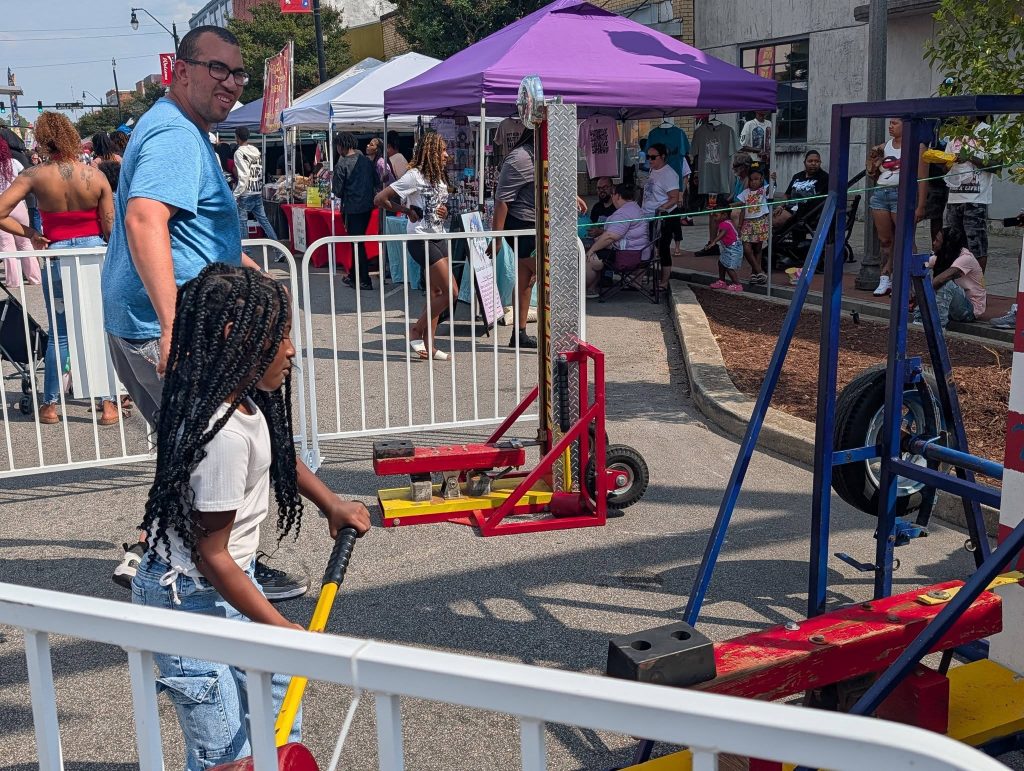 A girl plays a carnival strength test game as a man looks on. People shop at tents and booths in the background at an outdoor street fair.
