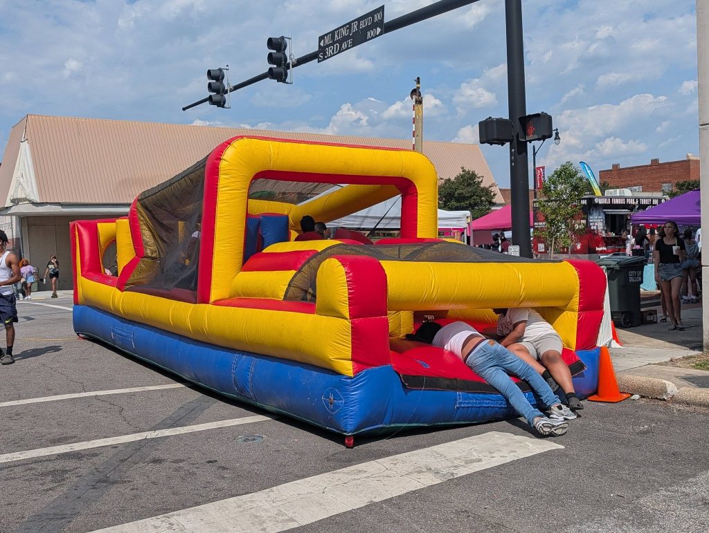 Two people crawl into a colorful inflatable obstacle course at a street festival, surrounded by tents, spectators, and street signs on a sunny day.