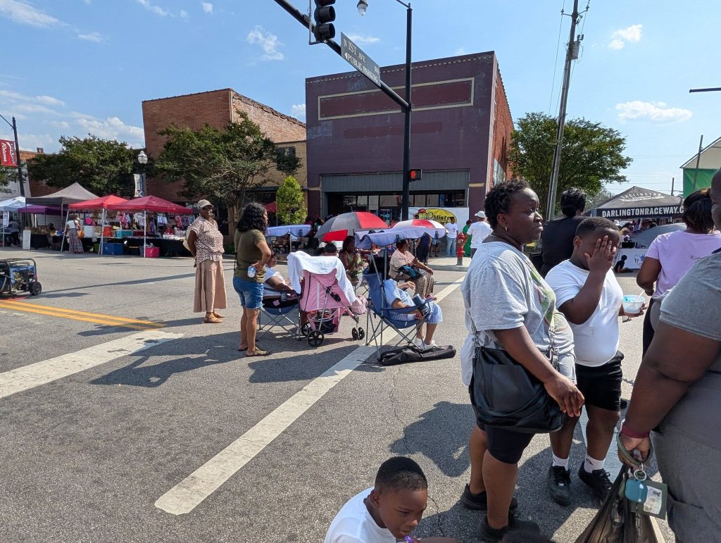 People gather on a sunny street for an outdoor event with tents, chairs, and vendors visible in the background.