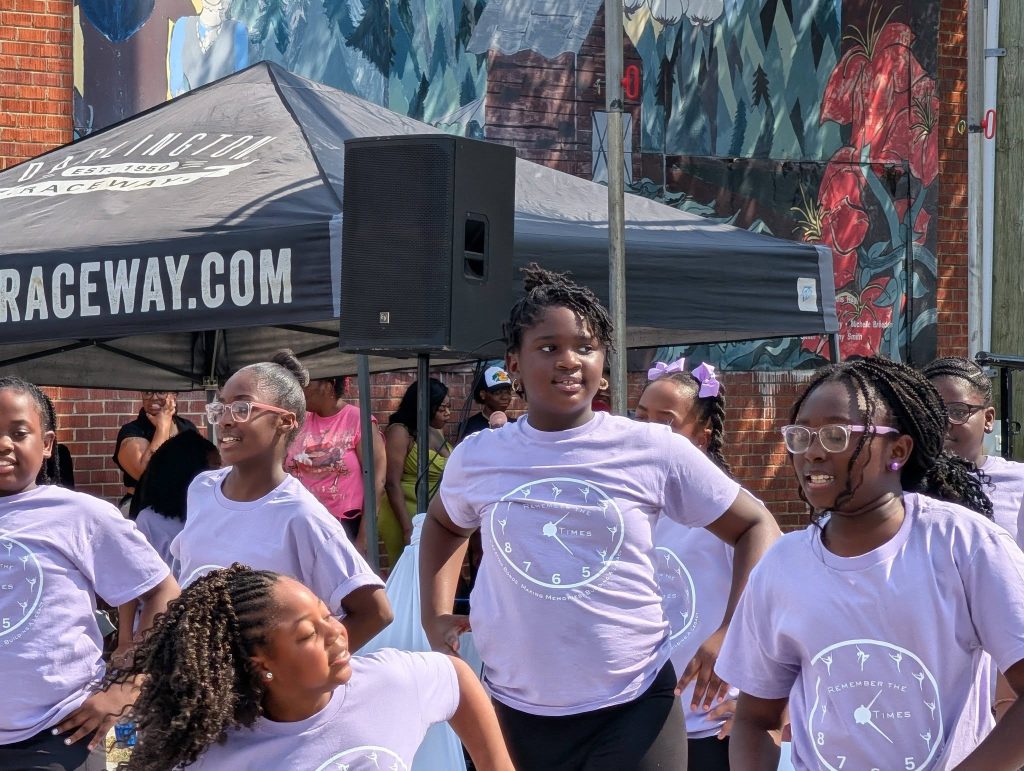 Group of young girls in matching light purple shirts performing a dance routine outdoors, with a crowd and colorful mural in the background.