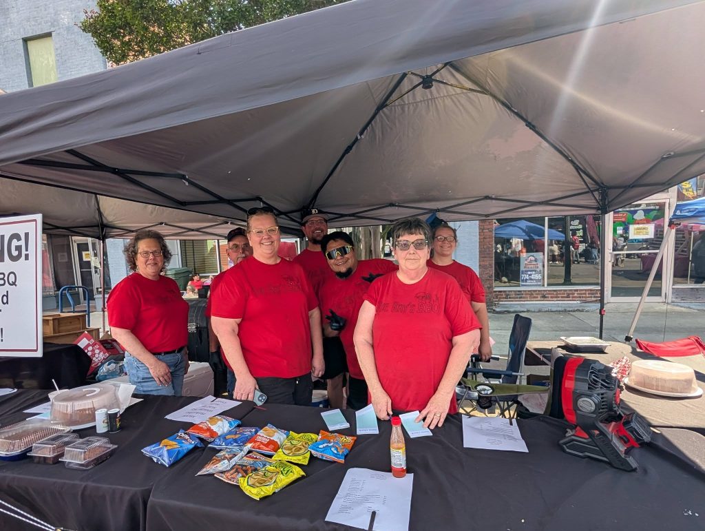 A group of people in matching red shirts stand under a canopy at an outdoor booth selling snacks and treats, smiling at the camera.