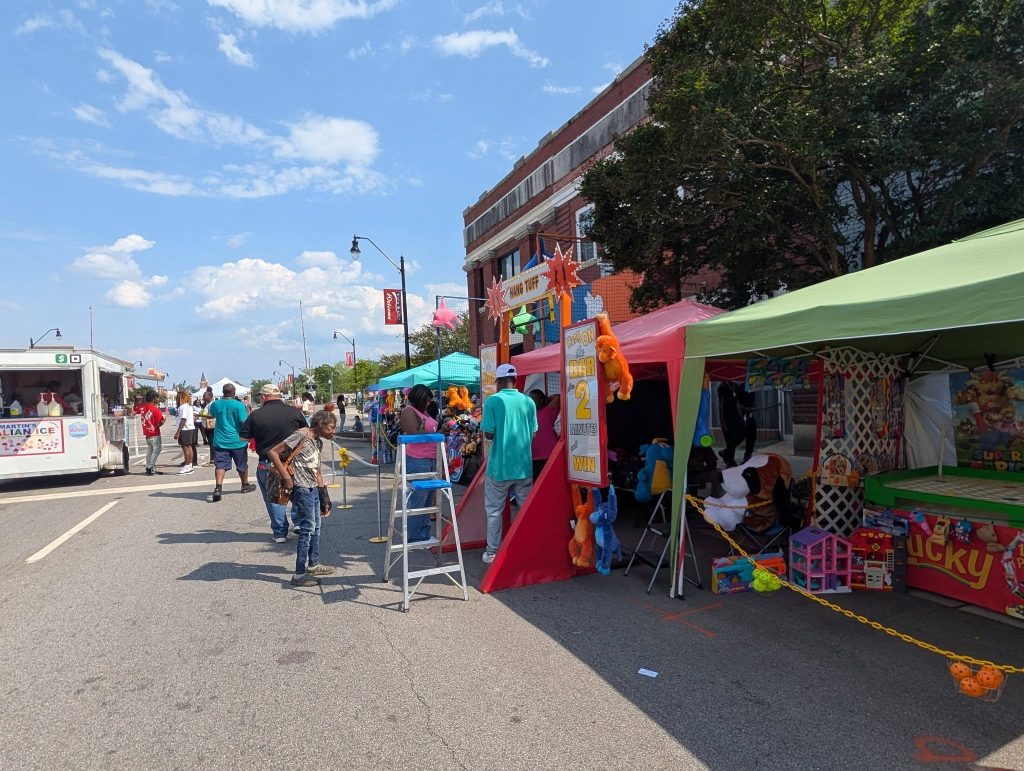 People attending a street fair with game booths, prizes, colorful tents, and a food truck under a bright blue sky on a sunny day.