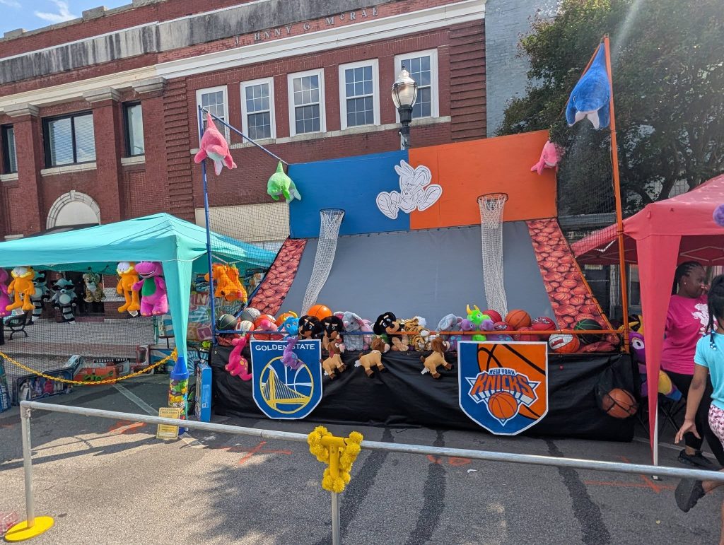 Outdoor basketball carnival game booth with stuffed animal prizes, basketballs, and team logos for Golden State Warriors and New York Knicks. Children and adults nearby.