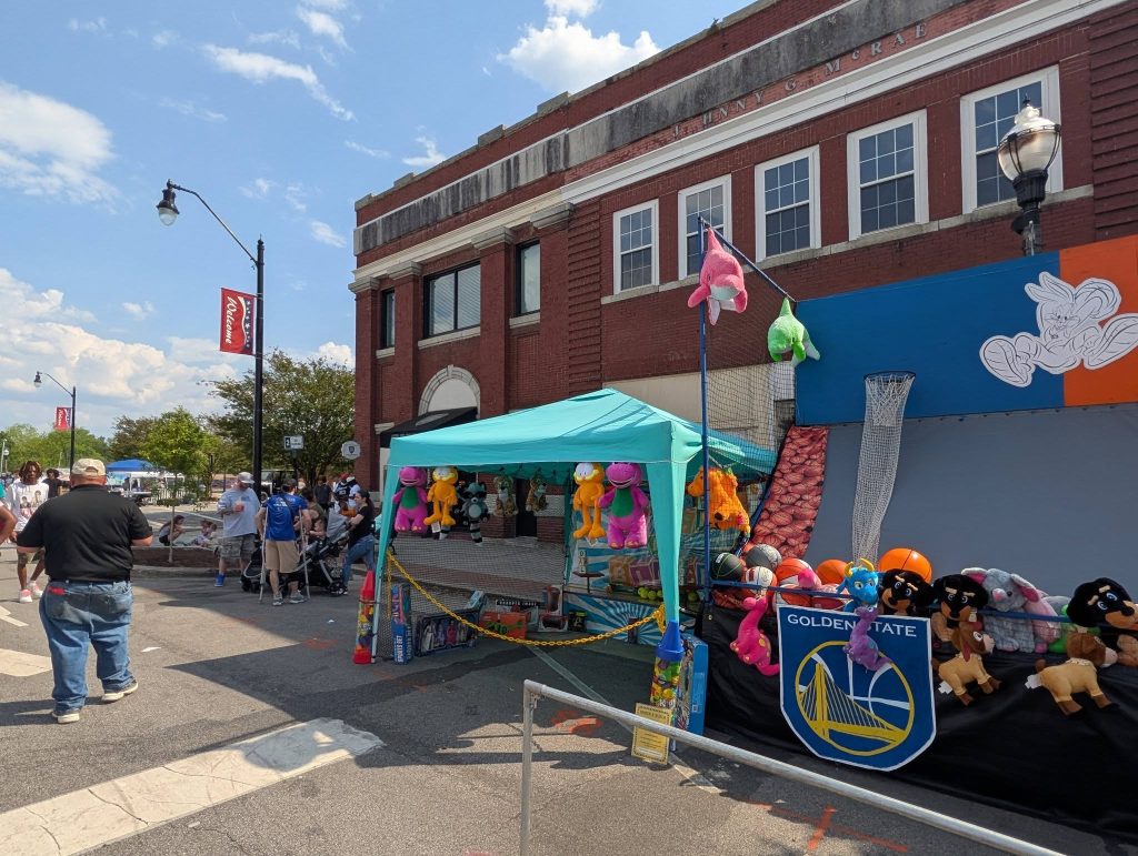 Outdoor street fair with people gathered around a basketball-themed carnival game booth with stuffed animal prizes and a building in the background.