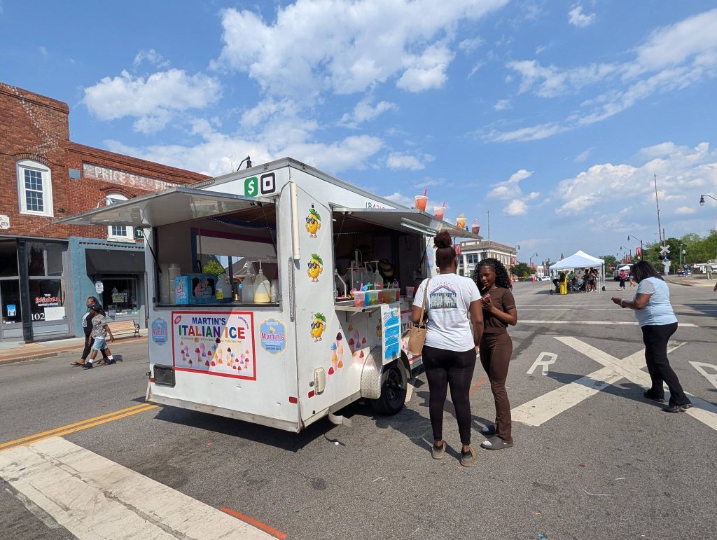 People gather at a street food truck labeled "Martin's Italian Ice" on a sunny day, with brick buildings and a vendor tent in the background.