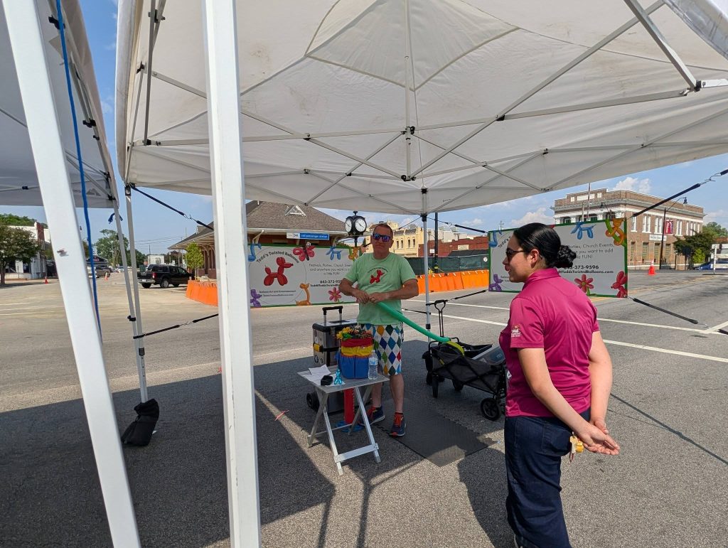 Two people under a white tent at a street fair; one shapes balloons, colorful decorations and event signs in the background.