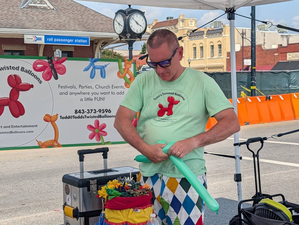 Man in sunglasses makes a balloon animal at an outdoor event booth, with colorful balloons and a sign advertising balloon art for parties behind him.