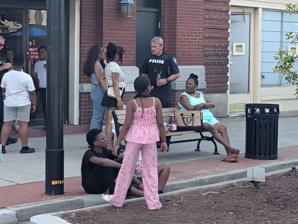 People gather on a city sidewalk; some are sitting, others standing, with a police officer and a woman seated on a bench in conversation.