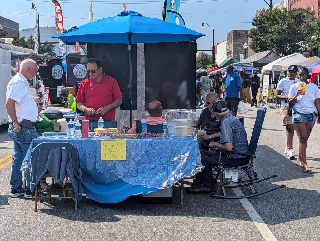 People gathered at a street fair booth under a blue umbrella, with a sign reading, "Let Me Tell You About My Jesus."