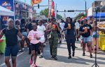 People walk through a lively outdoor street festival with food trucks, colorful tents, and drinks on a sunny day.