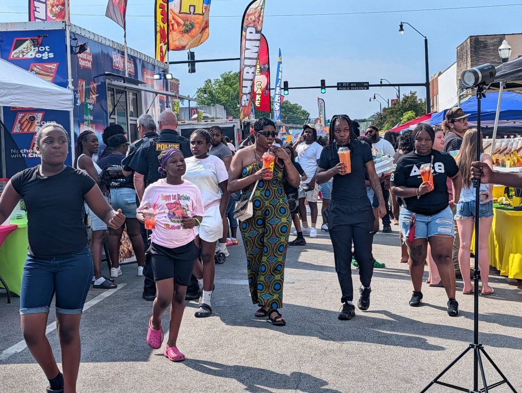 People walk through a lively street festival with food trucks, colorful flags, and drinks in hand on a sunny day.