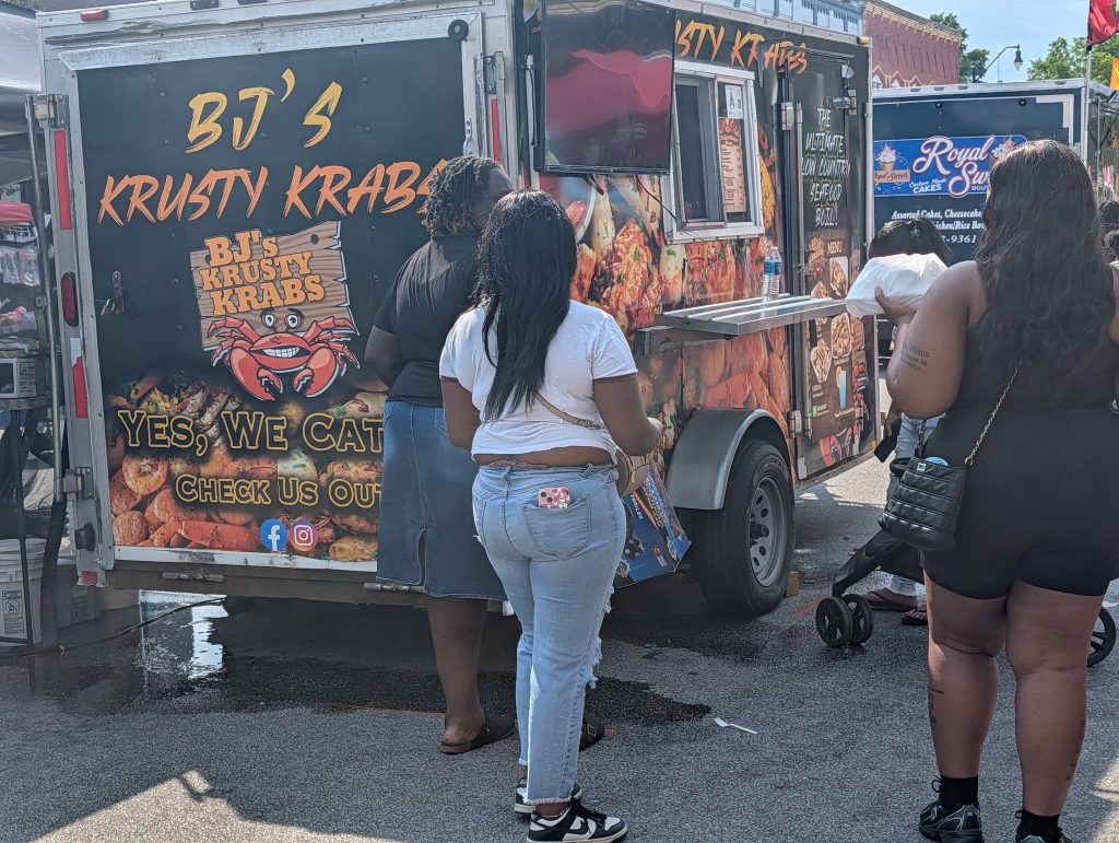 People standing in line at a BJ's Krusty Krabs seafood food truck at an outdoor event.