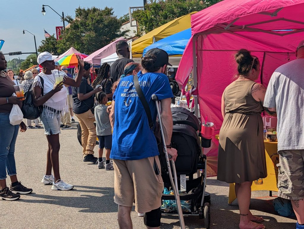 People gather at an outdoor street market with colorful tents, drinking beverages and shopping on a sunny day.